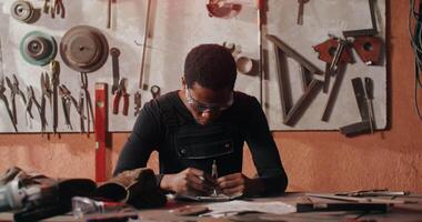 Work on creating sketch of future metal product in blacksmith's workshop. An African-American man dressed for work sketches while sitting at a desk in workshop full of tools. in 4k, red komodo video