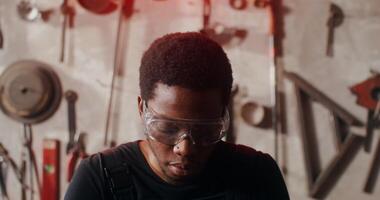 An African-American man dressed to work in a forge draws a sketch while sitting at a work table in a workshop full of tools, close-up. in 4k, red komodo video