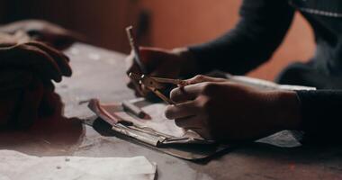 A young African-American man wearing goggles is working on a sketch while sitting at a work table in a blacksmith's workshop, close-up. in 4k, red komodo video