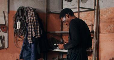 un' maniscalco nel occhiali usi un' digitale tavoletta per ricerca per idee per opera, seduta giù a il suo scrivania e di partenza per schizzo, Lavorando nel un' fucina tra un' varietà di Strumenti. nel 4k, rosso komodo video