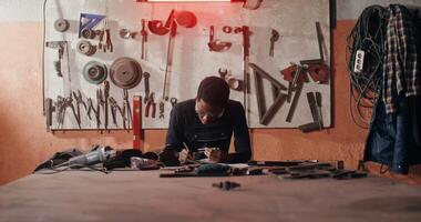 A young African-American blacksmith in goggles and work clothes draws a blueprint for a future product while sitting at a table in a workshop full of tools. in 4k, red komodo video