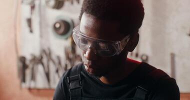 A young African-American man wearing goggles with a focused look works on sketching while sitting at a desk in a blacksmith's workshop full of tools. close-up. in 4k, red komodo video
