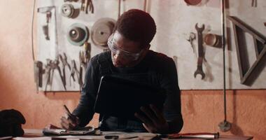 A young African-American man in protective transparent glasses with a focused look draws a sketch of a future product, looking in tablet, working in forge filled with tools. in 4k, red komodo video