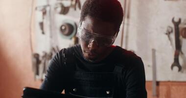 A young African-American man in goggles with a serious look draws a sketch of a product, using a digital tablet, working in a forge among a variety of tools. in 4k, red komodo video