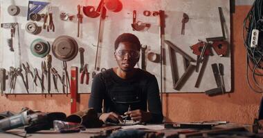 A young African American man in goggles and work clothes sits at a table in a workshop full of tools and smiles while looking at the camera. in 4k, red komodo video