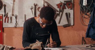 Work on creating a sketch of a future product in a blacksmith's workshop. An African-American man dressed for work sits at a desk in a workshop full of tools. in 4k, red komodo video