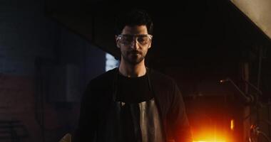A man in goggles and leather apron with sledgehammer in his hand looks seriously straight into the camera, standing in modern blacksmith's workshop against a kindled furnace. in 4k, red komodo video