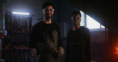 Two men, a Caucasian and an African American wearing goggles, dressed in work clothes are smiling looking directly at the camera while standing in a blacksmith's workshop. in 4k, red komodo video