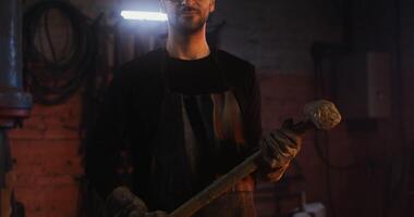 A young bearded man in goggles and a leather apron looks straight into the camera while standing in a modern blacksmith workshop with a sledgehammer in his hands, close-up. in 4k, red komodo video