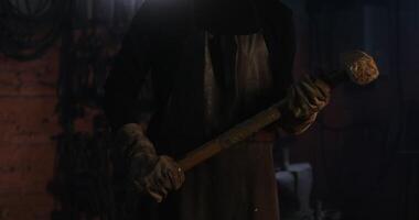 A young handsome man in a leather apron in goggles with a sledgehammer in his hands looks directly into the camera while standing in a modern blacksmith workshop. in 4k, red komodo video