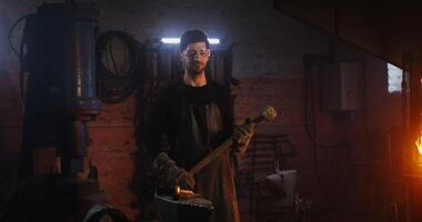 A young bearded man in goggles and a leather apron looks straight into the camera while standing in a modern blacksmith workshop with a sledgehammer in his hands. in 4k, red komodo video