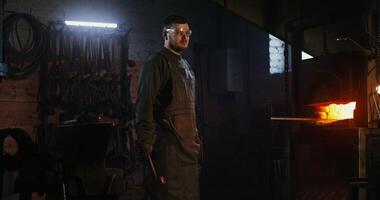 A blacksmith dressed in a hazmat suit turns his head and looks straight into the camera as he stands near a burning furnace while working on melting metal. 4k , red komodo video