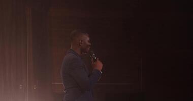 A handsome young African American man speaks into a microphone while standing on stage. filming under concert lighting. in 4k, red komodo video
