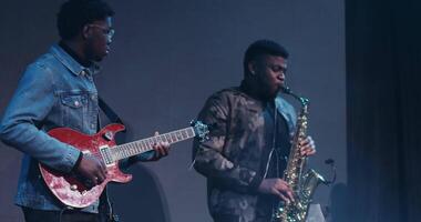 Two African American men play musical instruments while standing on the stage, close-up. Electric guitar and saxophone in the hands of musicians, concert lighting. in 4k, red komodo video