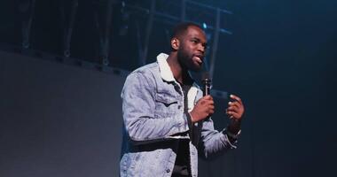 A handsome young African American man speaks into a microphone and actively gesticulates with his hands while standing on stage. filming under concert lighting. in 4k, red komodo video