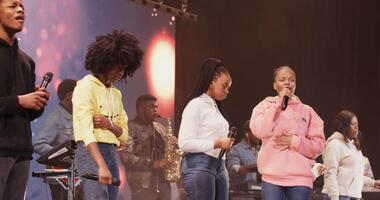 A musical group of young people of African American appearance sing into microphones and play musical instruments while standing on stage. Concert of a musical group. in 4k, red komodo video