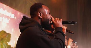 A young African-American man broke out in sweat while singing into a microphone as part of a band. filming from the side, concert lighting, stage performance. in 4k, red komodo video