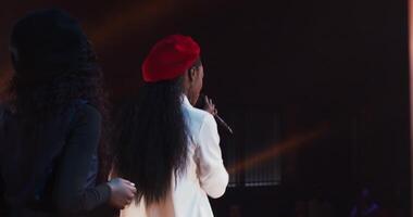 An African-American woman in a red beret sings into a microphone on stage during a concert as part of a musical group, from behind. 4k , red komodo video