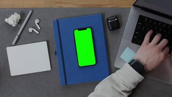 Close-up of male hands typing on a laptop keyboard, next to it lies a smarton with a green screen on a notepad. Mockup, chroma key, copy space video