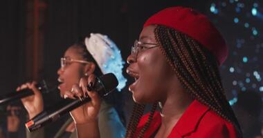 A young African-American woman with a lot of braids on her head in a beret sings emotionally into a microphone, participating in a concert as part of group, side view . 4k , red komodo video