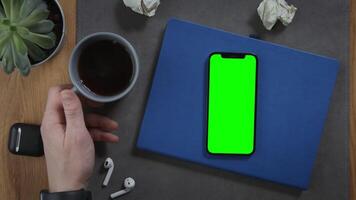 A man takes a cup of hot coffee, standing on desktop next to a mobile phone with a green screen, lying on a notebook with pieces of paper crumpled next to it, close-up. Mockup, chroma key, copy space video