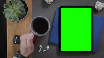 A man puts a cup of hot coffee on the desktop next to a digital tablet with a green screen lying on a notepad with crumpled pieces of paper nearby, close-up. Mockup, chroma key, copy space video