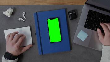A man is typing on a laptop keyboard while sitting at his desk with a green screen smartphone and notepad lying next to him, close-up. Mockup, chroma key, copy space video