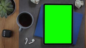 A man takes a cup of hot coffee on the desktop next to a digital tablet with a green screen lying on a notepad with crumpled pieces of paper nearby, close-up. Mockup, chroma key, copy space video