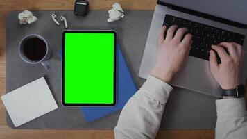 A digital tablet with a green screen lies on the desktop next to an open laptop, on the keyboard of which a man is typing, close-up, top view, no face. Mockup, chroma key, copy space video