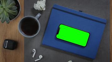 A mobile phone with a green screen lies on a notepad on the desktop, there is a cup of hot coffee nearby, close-up, no people. Mockup, chroma key, copy space video