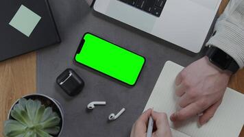 A man makes notes with his left hand in a notebook, lying next to a mobile phone with a green screen on the desktop, close-up of male hands. Mockup, chroma key, copy space video