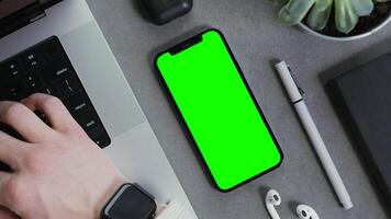Close-up of a mobile phone with a green screen lying on the desktop of a modern man typing on a laptop keyboard. Close-up of male hands, unrecognizable person. Mockup, chroma key, copy space video
