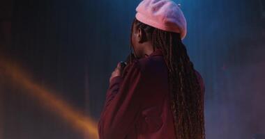 A young African-American woman with a lot of braids on her head in a beret sings emotionally into a microphone, participating in a concert as part of group, from the back. 4k , red komodo video