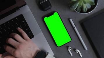 A mobile phone with a green screen lies on the desktop of a modern man typing on a laptop keyboard. Close-up of male hands. Mockup, chroma key, copy space video