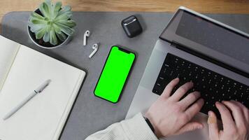 A stylish man is typing on a laptop while sitting at his desk, a smartphone with a green screen lies next to him. Close-up of male hands, unrecognizable person. Mockup, chroma key, copy space video