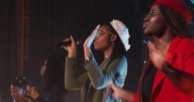 A group of young African-American women in berets with microphones in their hands clap their hands and begin to sing while standing on stage during a concert. 4k , red komodo video