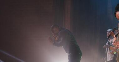 An African American man dances fervently and sings into a microphone while participating in a concert as part of a musical group. Incendiary dances, cheerful music. 4k , red komodo video