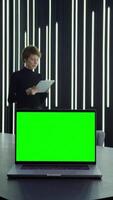 Young businesswoman is using a tablet while standing at her desk with an open green screen laptop standing on it, vertical . Chroma key, mockup, copy space. in 4k format, red komodo. video