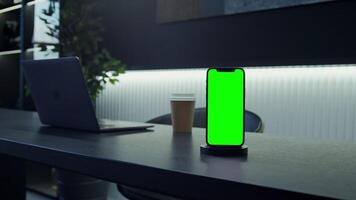 A mobile phone with a green screen stands on a stand on a desktop in front of an open laptop. Stylish office, desktop ready to work. Copy space, mocap, chroma key. in 4k, red komodo video