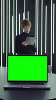 Businesswoman uses a tablet while standing near desktop with an open laptop with a green screen standing on it in a stylish office, vertical . Mockup, copy space. in 4k format, red komodo. video