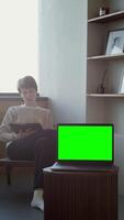 An open laptop with a green screen stands on a coffee table against the background of a young woman reading a book. Copy space, chroma key, mock up. 4k , red komodo, vertical video