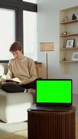 An open laptop with a green screen stands on a coffee table against the backdrop of a woman sitting on a sofa and reading a book. Copy space, chromakey, mock up. 4k , red komodo, vertical video