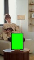 Young woman reading a book sitting on a sofa in front of a green screen tablet standing on a coffee table, copy space. 4k , red komodo, vertical video