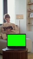 A young woman is reading a book sitting on a sofa in front of an open laptop with a green screen standing on a coffee table, copy space. 4k , red komodo, vertical video