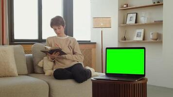 An open laptop with a green screen stands on a coffee table against the background of a woman reading a book, copy space, chroma key. in 4k format, red komodo. video