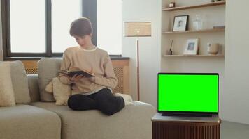 An open laptop with green screen stands on a coffee table against the background of a woman reading a book, sitting on a sofa in home interior. Chroma key, copy space. in 4k format, red komodo. video