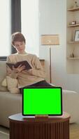 A tablet with a green screen stands on a coffee table against the background of a reading a book sitting on the sofa woman, copy space, chroma key. 4k , red komodo, vertical video