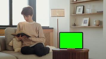 A tablet with a green screen stands on a table against the background of a woman reading a book, sitting on a sofa in a home interior. Chroma key, mock up, copy space. in 4k format, red komodo. video