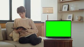 An open green screen laptop sits on a coffee table in front of a woman reading a book sitting in a stylish home interior. Chromakey, mockup. in 4k format, red komodo. video