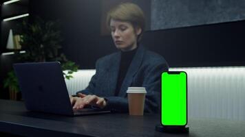 A mobile phone with a green screen stands on a stand on a desktop in front of a businesswoman working at a computer in a stylish office. Copy space, chromakey, mockup. in 4k format, red komodo. video